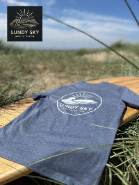 Blue t-shirt with 'Lundy Sky' logo on a wooden surface with grass and sky in the background